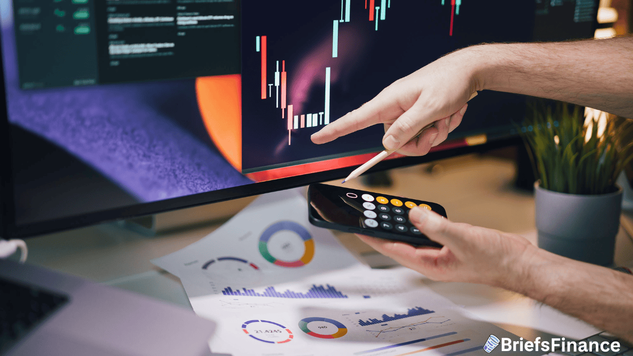 Person analyzing stock market charts on a computer monitor, using a smartphone calculator, with financial documents and graphs on a desk.