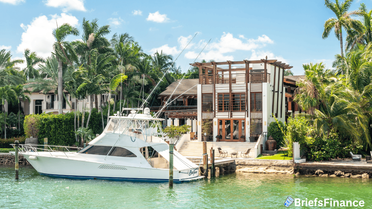 A white yacht is docked in front of a large modern waterfront house with palm trees and patio seating under a blue sky.