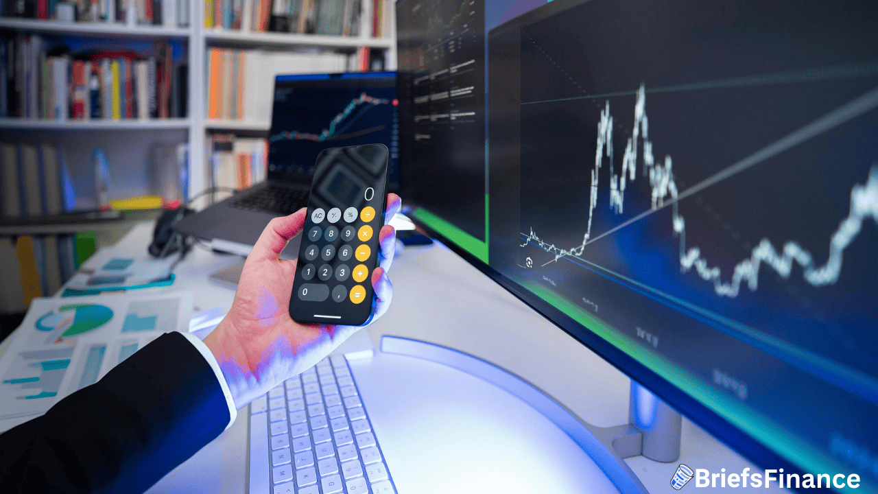 A person uses a calculator on a smartphone at a desk with financial charts displayed on monitors and documents nearby.