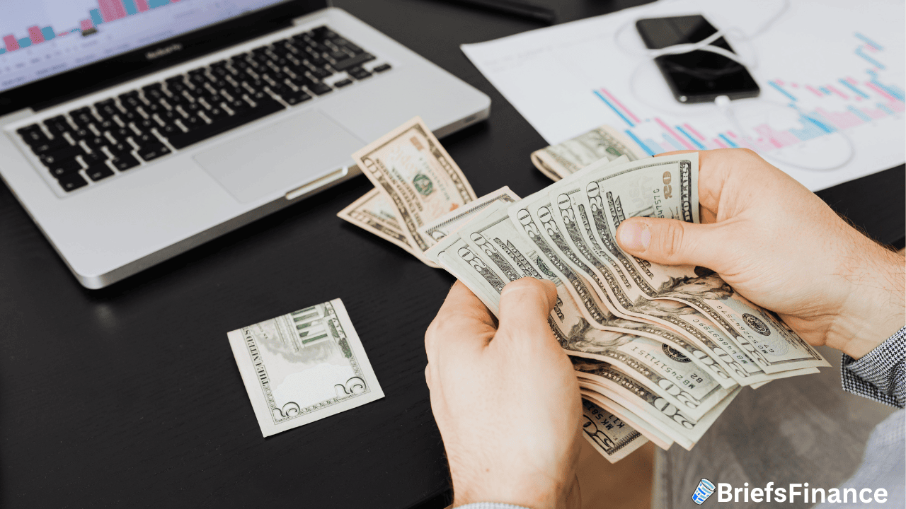 Person counting U.S. dollar bills at a desk with a laptop, smartphone, and financial charts in the background. "BriefsFinance" logo in the lower right corner.
