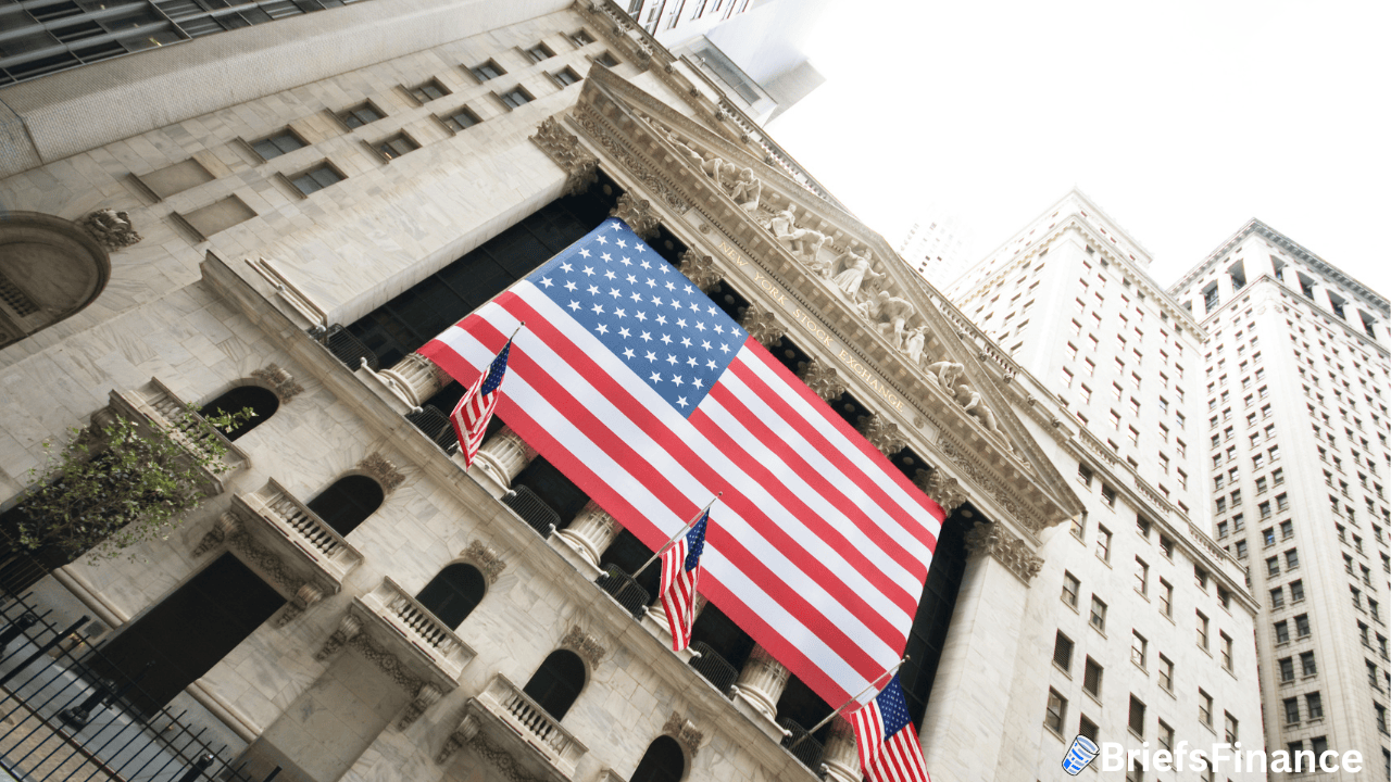 The front of the New York Stock Exchange building is draped with a large American flag and several smaller flags, surrounded by tall buildings.