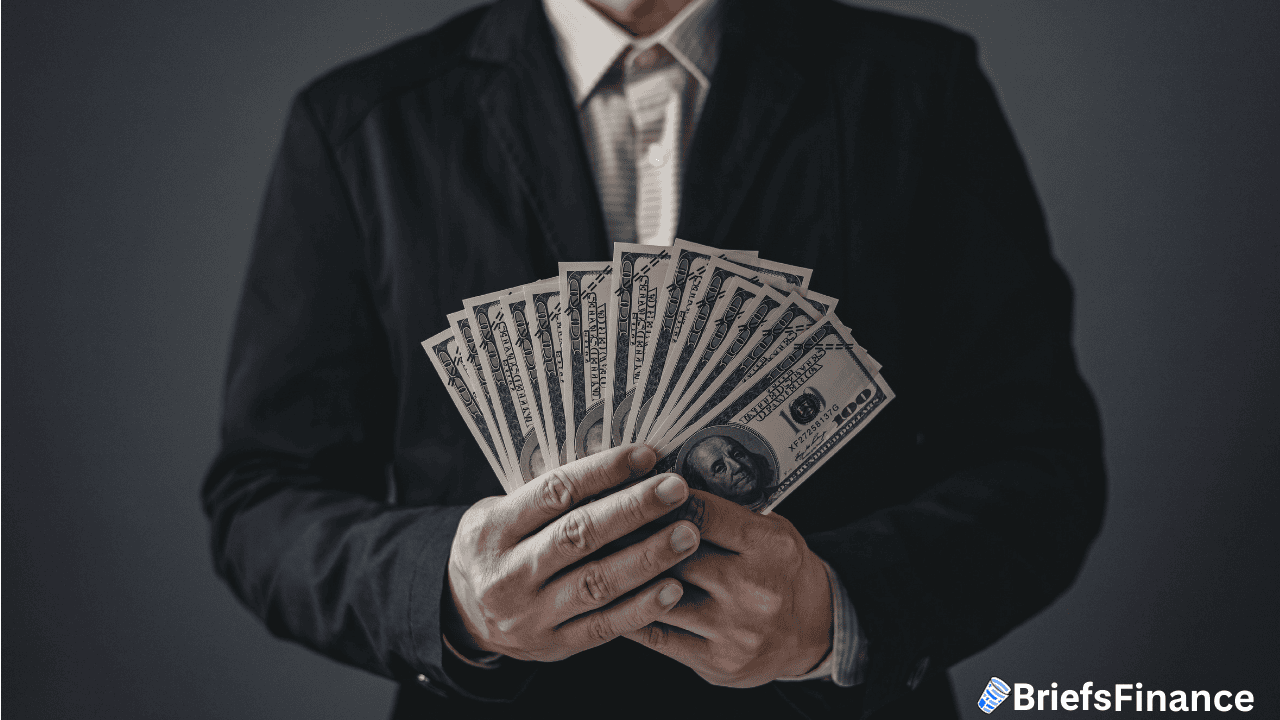 A person in a dark suit holds a fanned-out stack of US hundred-dollar bills; BriefsFinance logo appears in the bottom right corner.