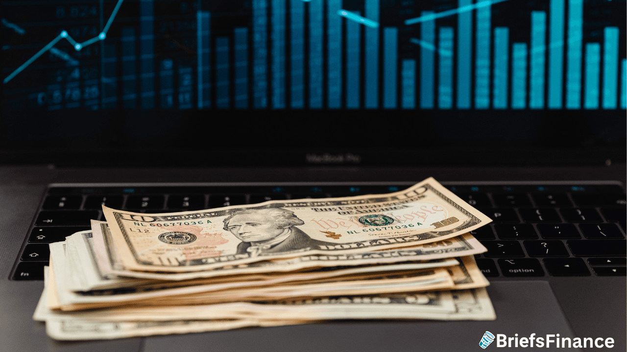 A stack of US dollar bills on a laptop keyboard, with financial graphs displayed on the screen in the background.