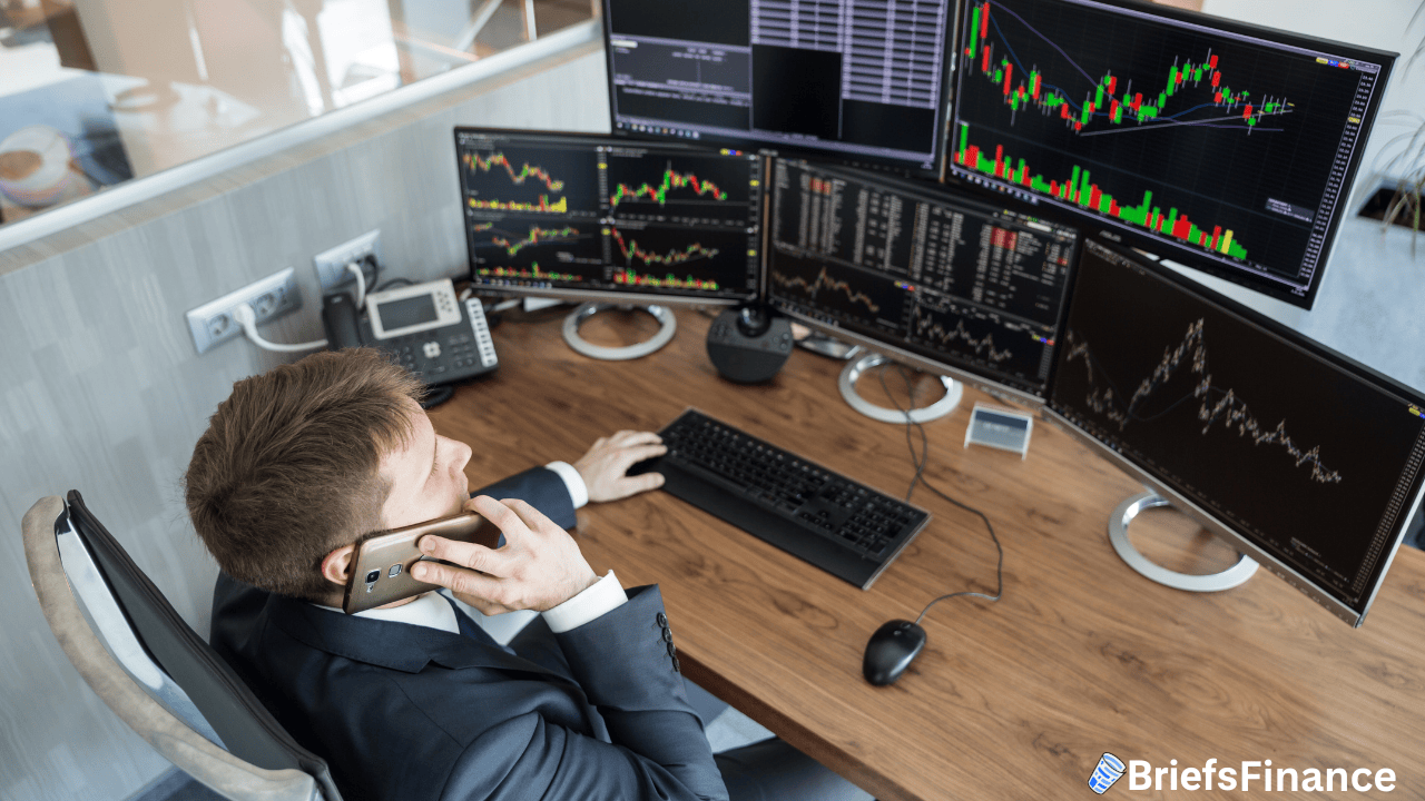 A man in a suit sits at a desk with multiple monitors displaying financial charts, talking on the phone in an office setting.