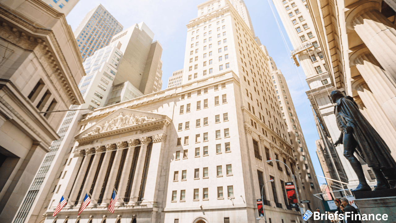View of tall financial district buildings with the New York Stock Exchange, American flags, and a statue in the foreground under a clear sky.
