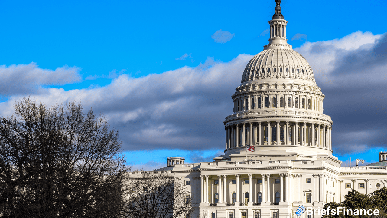 The United States Capitol building stands against a partly cloudy blue sky, with trees in the foreground and the BriefsFinance logo in the corner—a fitting scene as Polymarket and Kalshi debates intensify amid talks of a Senate trading ban.
