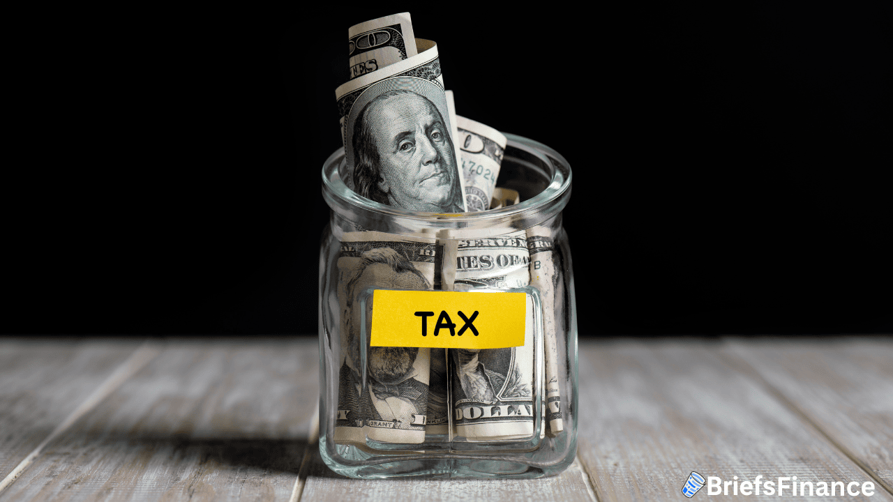 A glass jar labeled "TAX" filled with rolled U.S. dollar bills sits on a wooden surface against a black background.