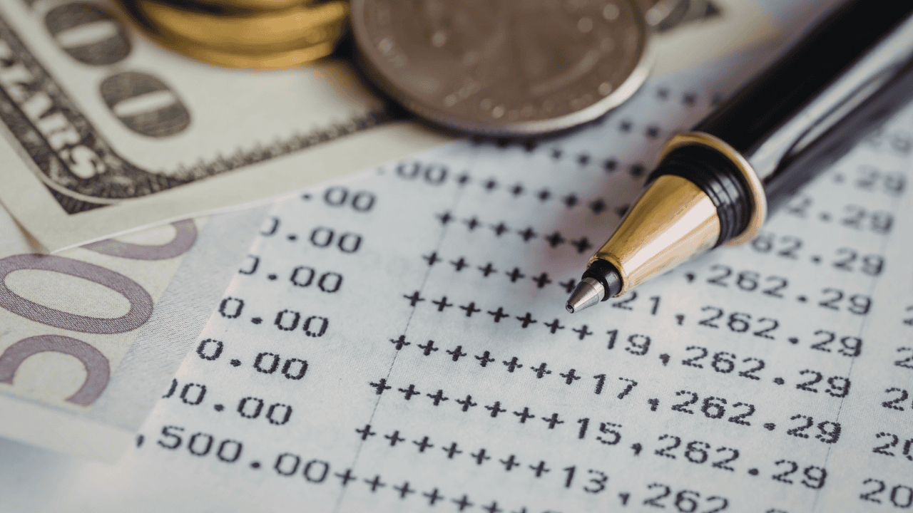 A pen rests on a balance sheet beside paper currency and coins, ready for an investor to read a balance sheet and analyze financial details.