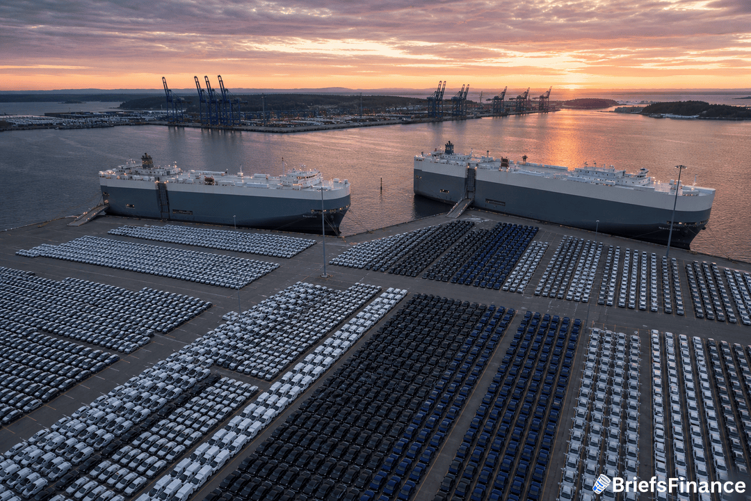 Rows of parked cars are lined up at a port as two large cargo ships are docked nearby under a sunset sky.